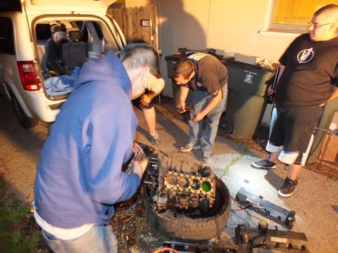 2003 Ford Crown Victoria - worker handles an oily engine block on a tire while others use a flashlight in the dark