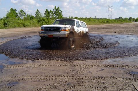 1995 Ford Bronco - A white full-size Ford Bronco charges front-first through a muddy water hole, throwing brown spray off its front tire. It sits lifted on of…