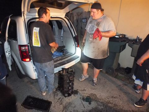 2003 Ford Crown Victoria - man in a do-rag and another work near a silver minivan with an engine block on the pavement