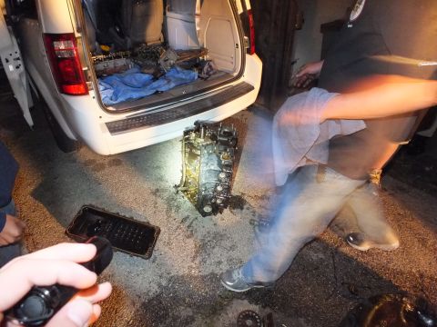 2003 Ford Crown Victoria - Worker steps past an engine block and oil pan behind a silver minivan's open rear hatch