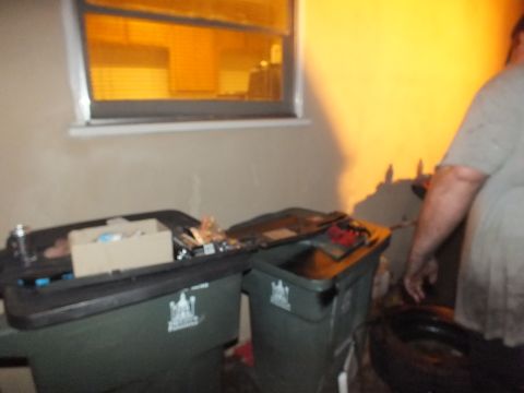 2003 Ford Crown Victoria - Person standing beside green roll-out trash bins topped with tools and cardboard under orange-lit garage wall