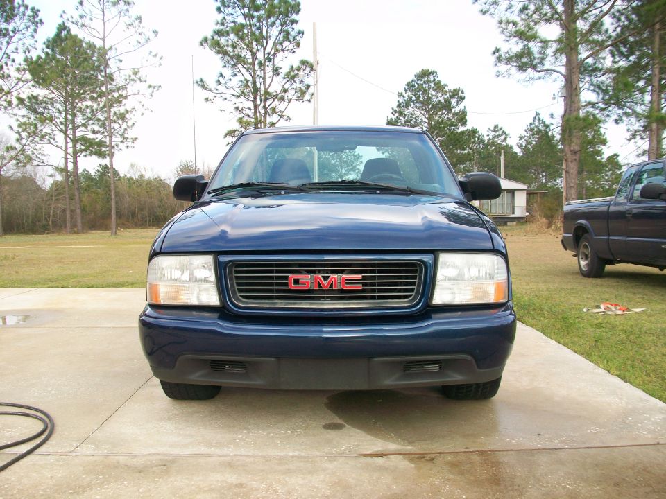 Front view of a blue 2000 GMC Sonoma pickup truck parked on a concrete driveway. Another blue pickup truck is visible in the background