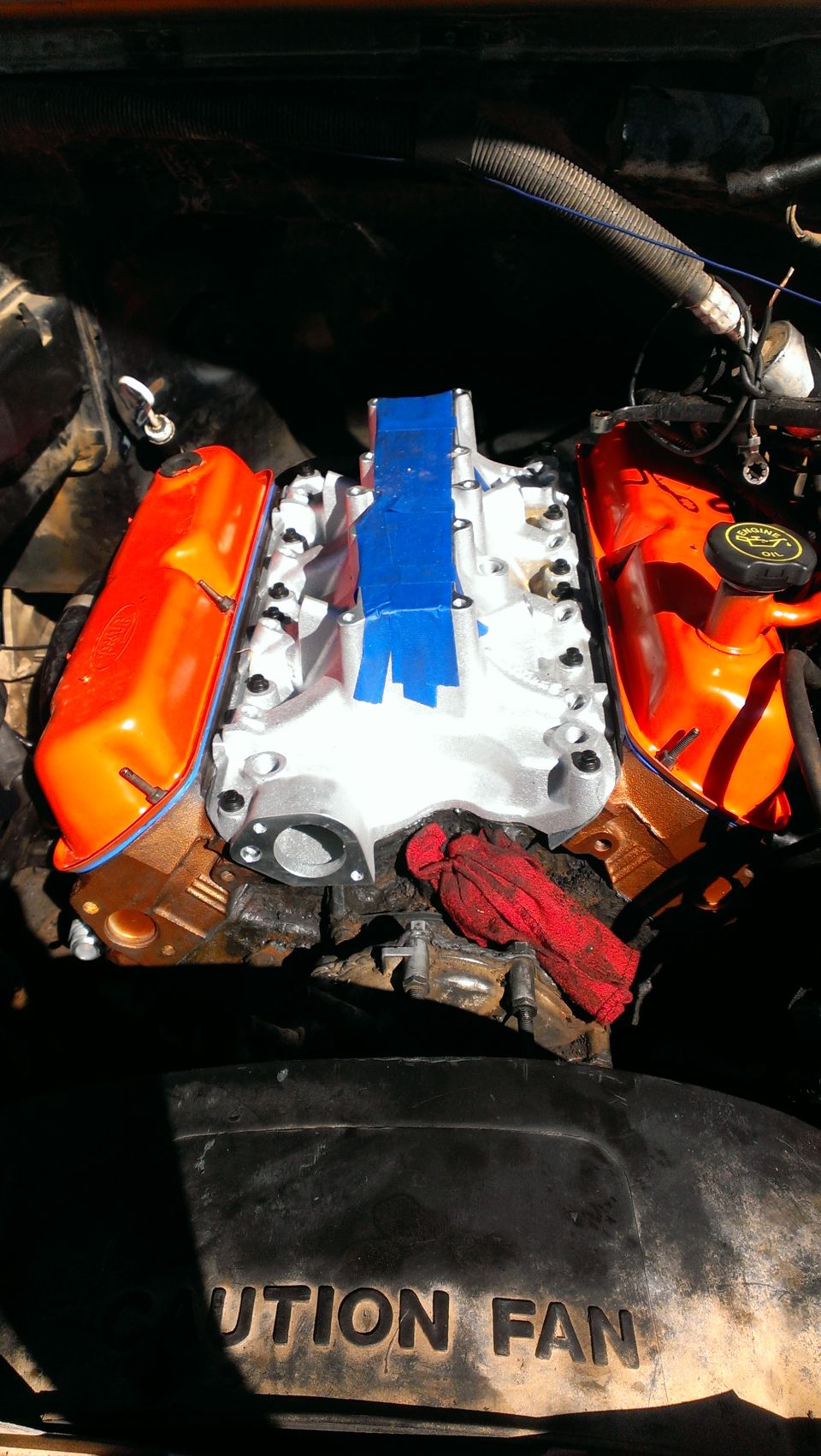 Engine with orange valve covers and a silver intake manifold taped with blue tape. A red rag is on the engine