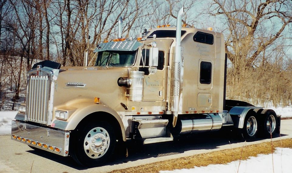 Gold Kenworth semi tractor with sleeper cab and chrome stacks, parked near bare trees and snow