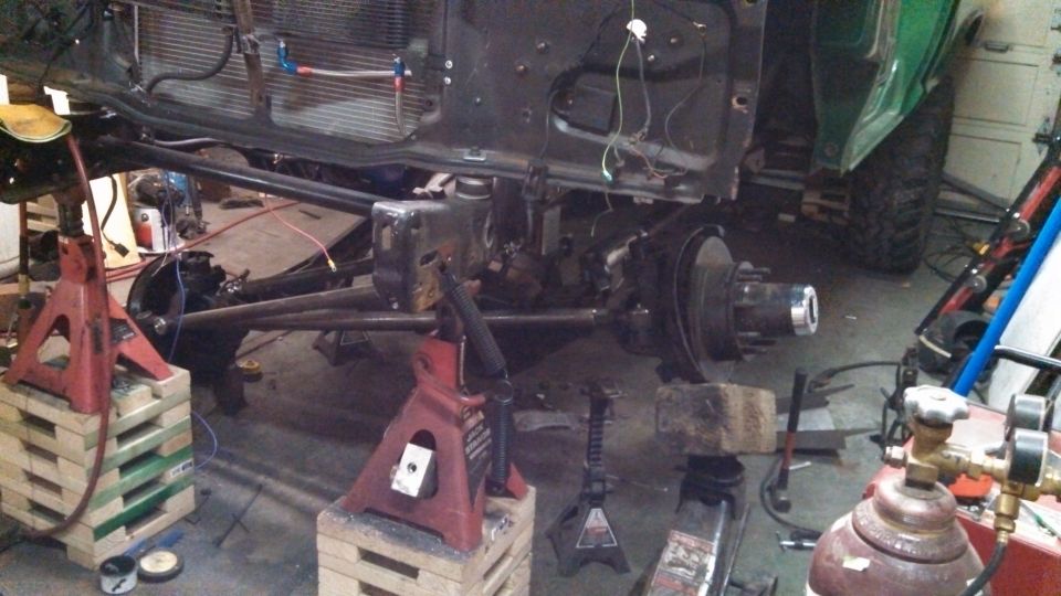 Vehicle frame and suspension components on jack stands in a workshop. Part of a green vehicle is visible on the right