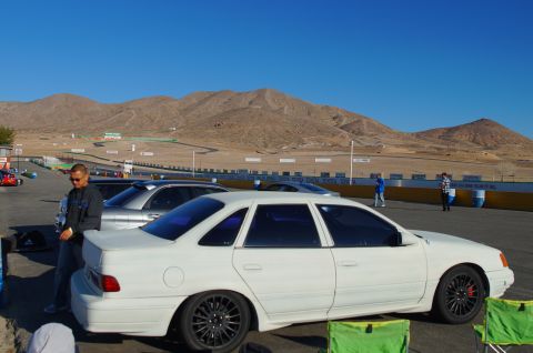1995 Ford Taurus - A white 1995 Ford Taurus is parked at the edge of a race track with mountains in the background.