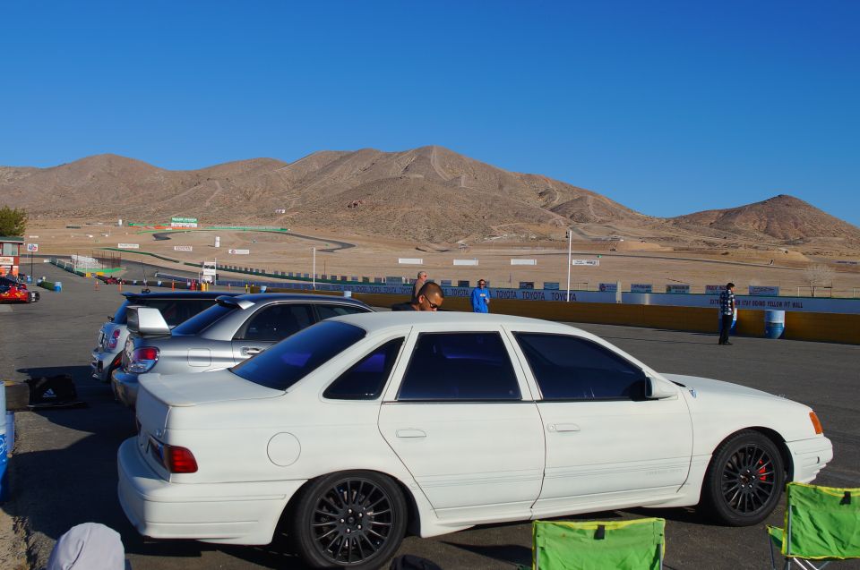 White 1995 Ford Taurus parked on asphalt with a racetrack and mountains in the background. Another silver car is parked in front of it