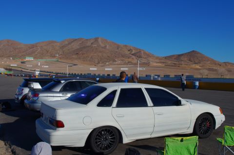 1995 Ford Taurus - A white sedan sits on the asphalt at a race track, with other vehicles and desert mountains in the distance.