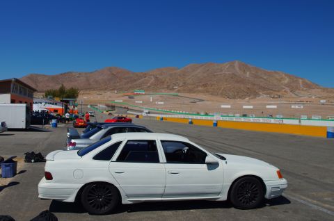 1995 Ford Taurus - A white 1995 Ford Taurus is parked on the asphalt near a race track with other cars and mountains visible.