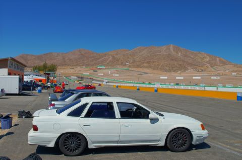 1995 Ford Taurus - A white 1995 Ford Taurus sedan is parked at a race track with mountains in the background.