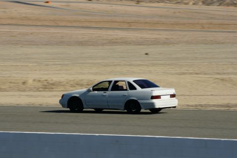 1995 Ford Taurus - A white sedan is captured in motion on a paved track with a dirt infield and distant cones.