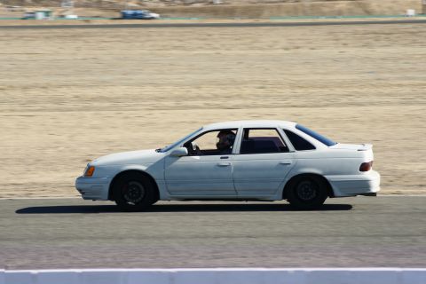 1995 Ford Taurus - A white sedan, likely a 1992 model, drives on a race track with a driver wearing a helmet.