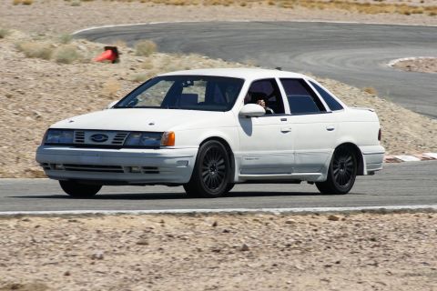 1995 Ford Taurus - A white 1995 Ford Taurus sedan navigates a turn on a race track, its sporty stance and black aftermarket wheels.