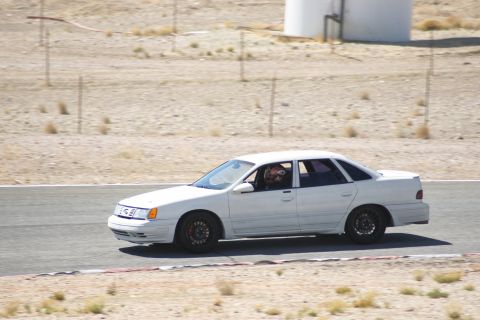 1995 Ford Taurus - White 1995 Ford Taurus sedan driving on a race track with a driver wearing a helmet.