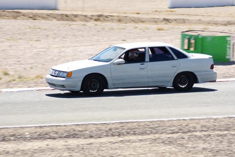 1995 Ford Taurus - A white sedan with a driver wearing a helmet is on a race track, possibly during a track day event.