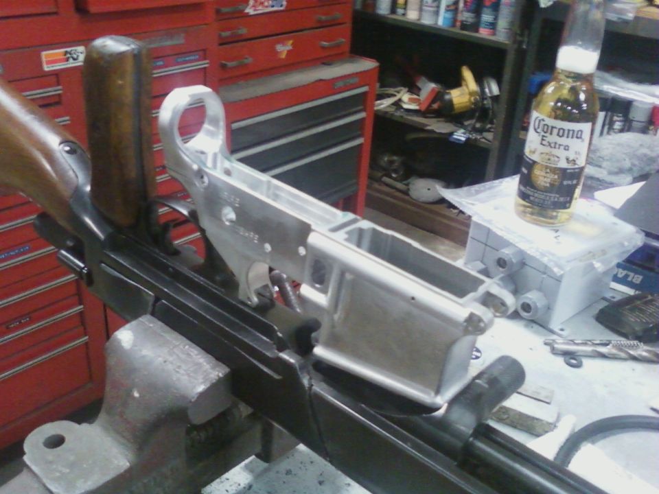Aluminum gun receiver held in a vise next to a wooden stock and a bottle of Corona Extra