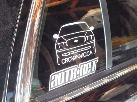 2003 Ford Crown Victoria - White CrownVic.ca outline and ADTR.net logo decals on a tinted side window with a faint round sticker behind