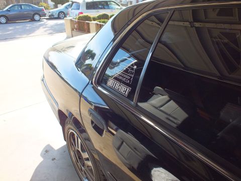 2003 Ford Crown Victoria - Glossy black sedan rear quarter showing CrownVic.ca and ADTR.net decals, alloy wheel and parked cars behind