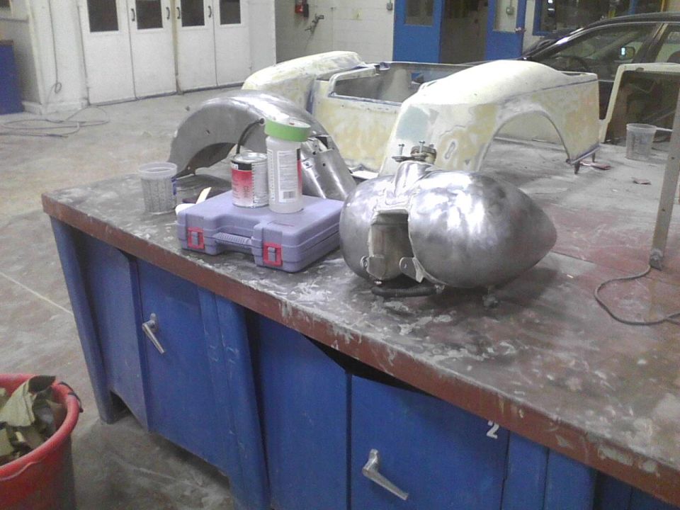 Metal car body parts, including a fuel tank and fender, on a blue workbench with paint cans and a toolbox