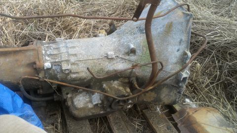 1995 Ford Bronco - A weathered transmission part with rusty metal lines sits on dry grass.
