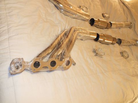 2003 Ford Crown Victoria - New plastic-wrapped tubular exhaust header beside two catalytic converters laid on a cream bedspread