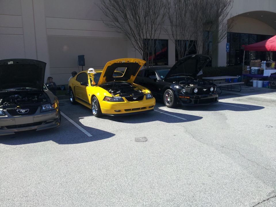 Three Ford Mustangs parked in a lot, hoods up. One is yellow, one is black, and one is grey