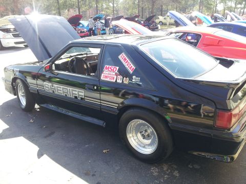 1965 Ford Mustang - A black Ford Mustang, modified for racing, displays "SHERIFF" text and various decals on its side.