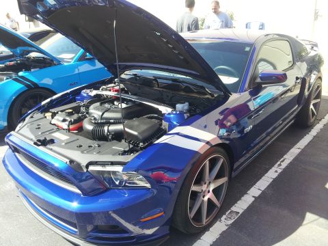 1965 Ford Mustang - A blue Ford Mustang with its hood open is parked next to another blue Mustang, displaying its engine and white racing stripes.