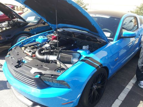 1965 Ford Mustang - Blue Ford Mustang with black racing stripes and hood open, revealing the engine bay. Another car is visible in the background.