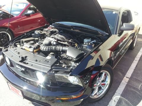 1965 Ford Mustang - A black Ford Mustang is parked with its hood open, revealing the engine bay and chrome wheels, with another red Mustang in the background.