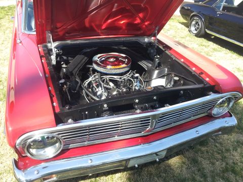 1965 Ford Mustang - Engine bay of a red classic car, with a chrome air cleaner and visible engine components. A second classic car is parked nearby.