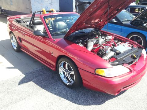 1965 Ford Mustang - A red convertible with its hood open, revealing the engine bay. Another car is visible to the right.