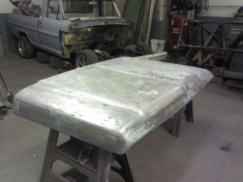 1967 Ford F-100 - A bare metal truck hood rests on a stand, with a partially disassembled truck visible in the workshop behind it.