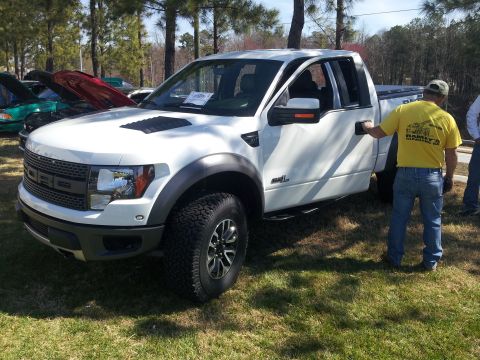 1965 Ford Mustang - A white Ford F-150 SVT Raptor pickup truck is displayed at an outdoor event, with other vehicles visible in the background.