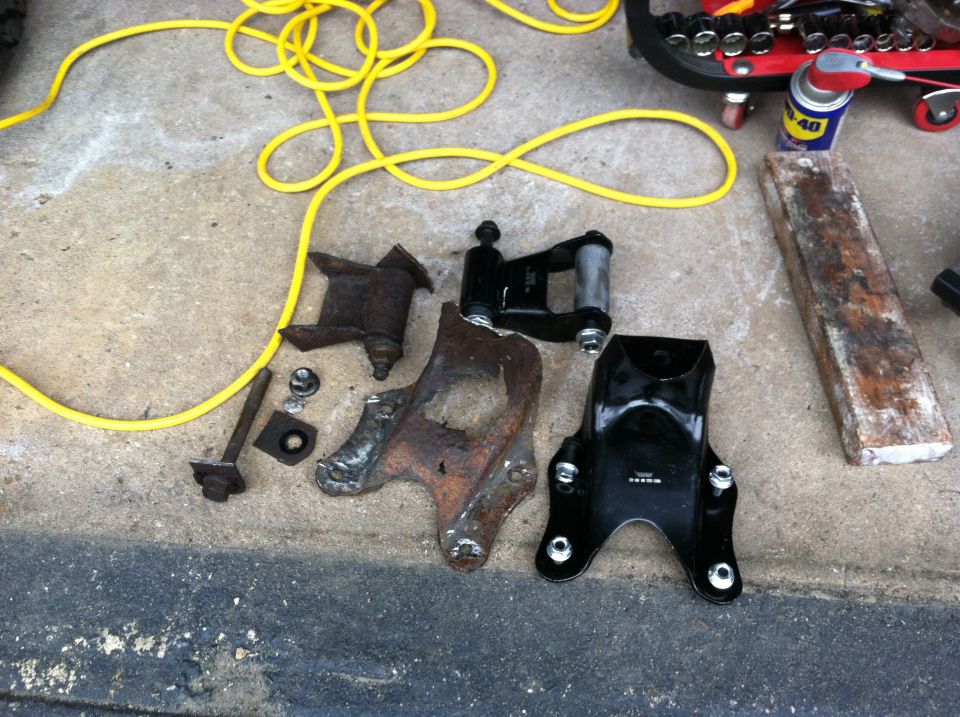 Several vehicle suspension parts, some rusty and some new black ones, laid out on concrete with a yellow cord and tools nearby