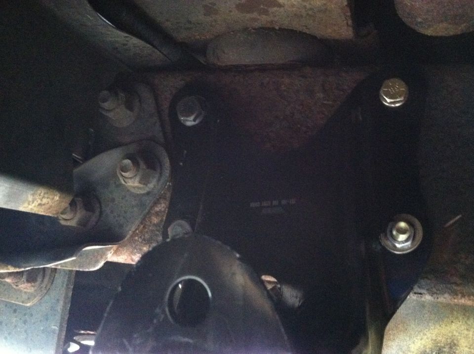 Close-up of a black metal bracket bolted to a rusty vehicle frame, with visible bolts and suspension parts