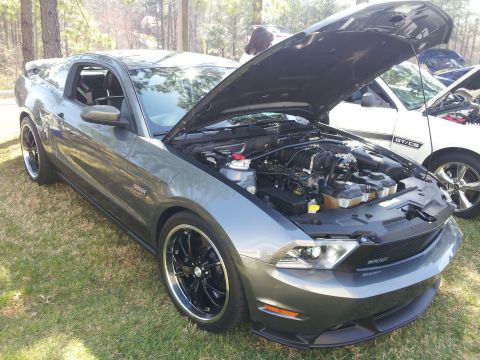 1965 Ford Mustang - A gray Ford Mustang is parked on grass with its hood open, revealing the engine bay. A white car is visible to the right.