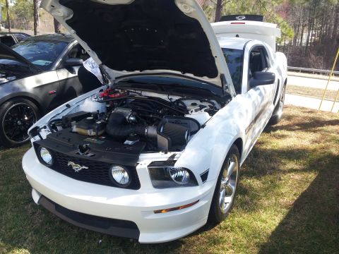 1965 Ford Mustang - A white Ford Mustang from the mid-2000s is displayed with its hood open, parked outdoors.