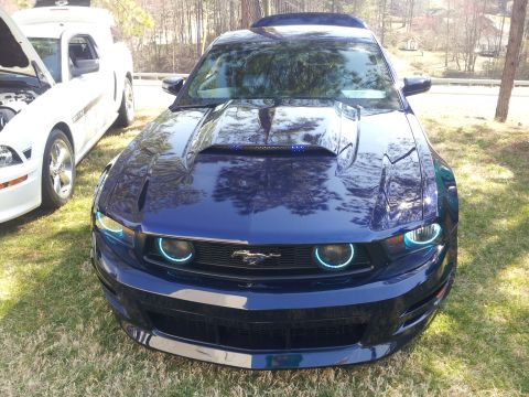 1965 Ford Mustang - A blue Ford Mustang with a prominent hood scoop and distinctive halo headlights is shown parked next to another Mustang.