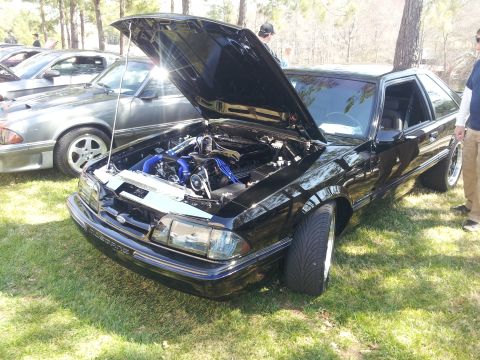 1965 Ford Mustang - A black Mustang with its hood up, revealing the engine bay, parked on grass next to another vehicle.