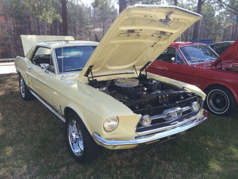1965 Ford Mustang - A yellow 1967 Ford Mustang coupe is displayed with its hood open, revealing the engine bay. Another Mustang is visible behind it.