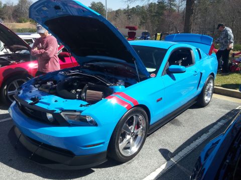 1965 Ford Mustang - A bright blue Ford Mustang with racing stripes is displayed with its hood and trunk open at an outdoor car show.