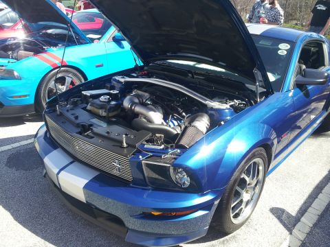 1965 Ford Mustang - Blue Ford Mustang with racing stripes and hood open, revealing its engine bay. Another Mustang is parked nearby.