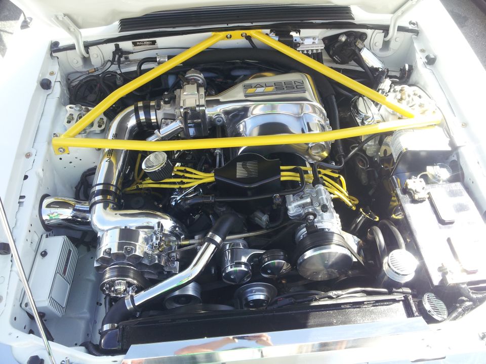Engine bay with a chrome supercharger and yellow spark plug wires, under a yellow strut bar