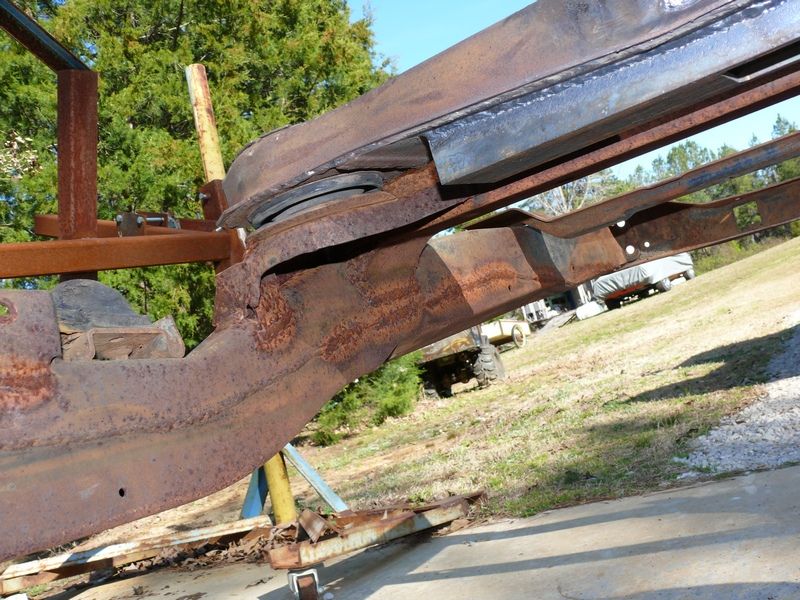 Rusted vehicle frame on stands, with a covered vehicle and tractor in the background
