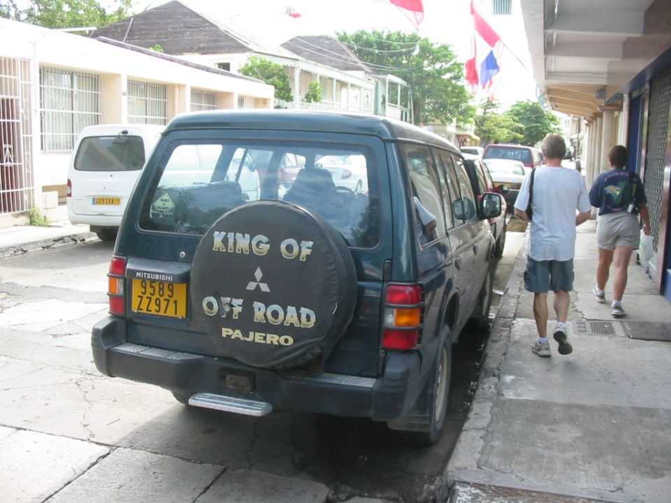 Mitsubishi Pajero parked on street, spare tire cover says 'KING OF OFF ROAD'. White van parked behind it