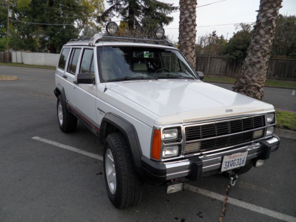 White Jeep Cherokee with roof rack and lights. License plate reads 3XQG316
