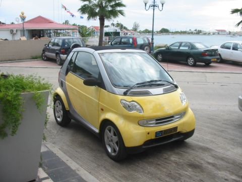 1992 Ford Bronco - A yellow Smart Fortwo is parked on a paved area near a body of water, with several other vehicles visible in the background.