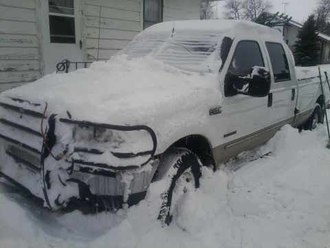 2000 Ford F-250 - A white Ford F-250 pickup truck is heavily covered in snow, with only parts of the grille guard and tires visible.