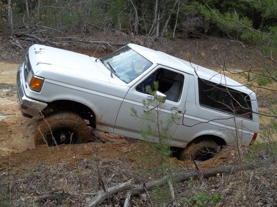 White 1987 Ford Bronco stuck in mud, front right wheel deep in dirt, off-road scene